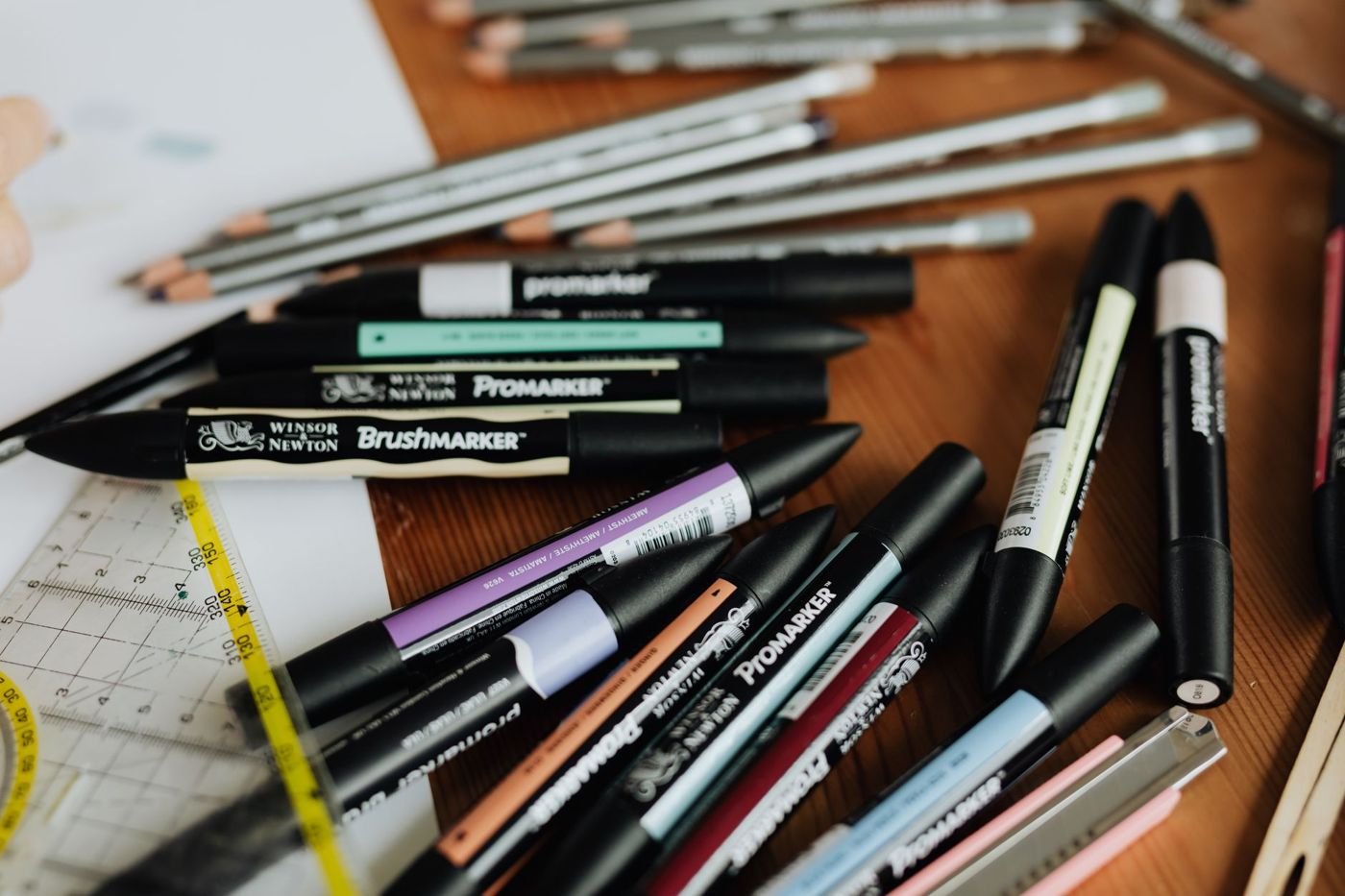Rotuladores Promarker de doble punta en tonos pastel y oscuros y lápices mezclados sobre una mesa de madera, junto a una regla