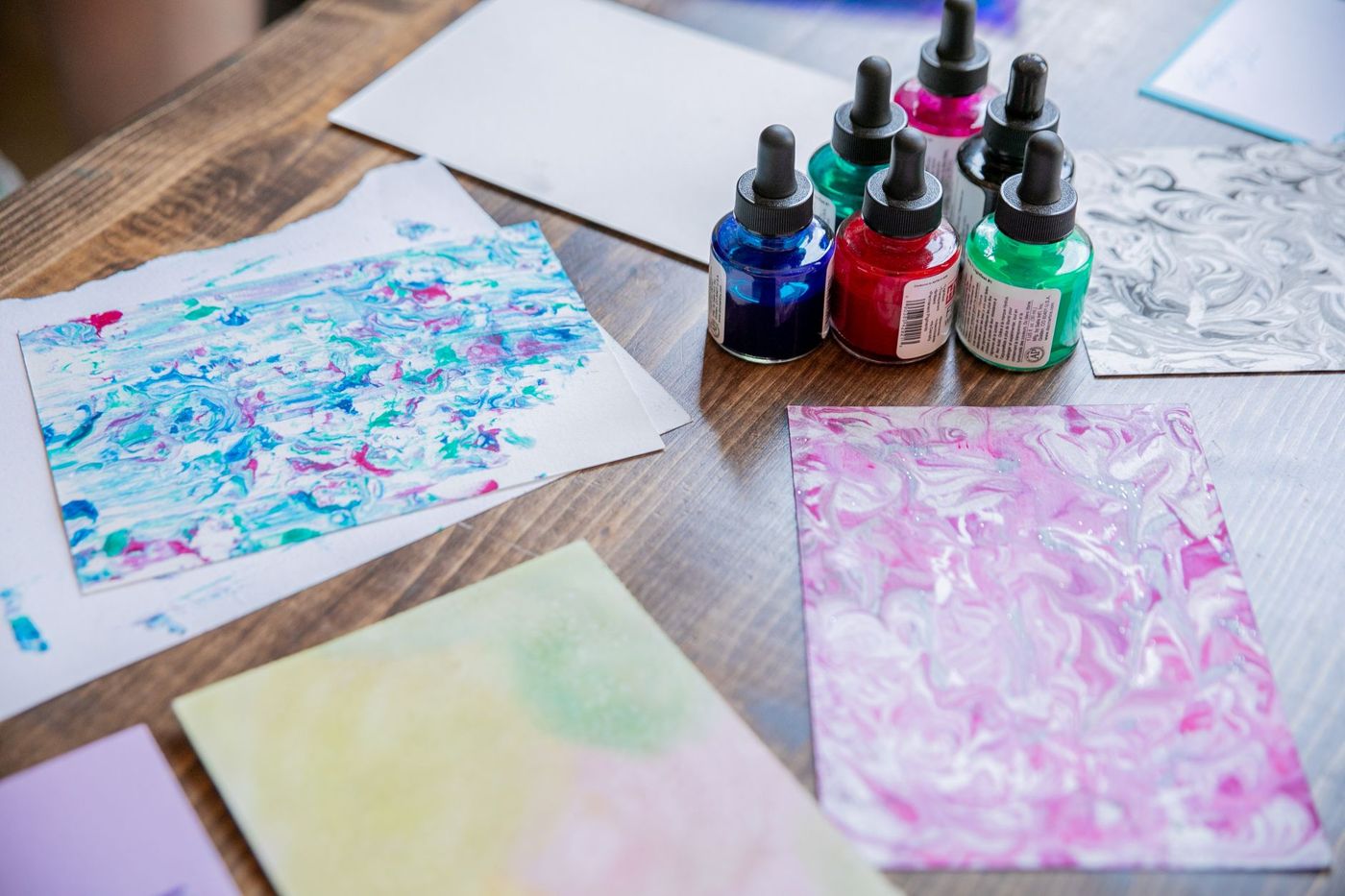 Six dropper bottles of ink in blue, red, green and black next to marbled papers with pink and blue gradients