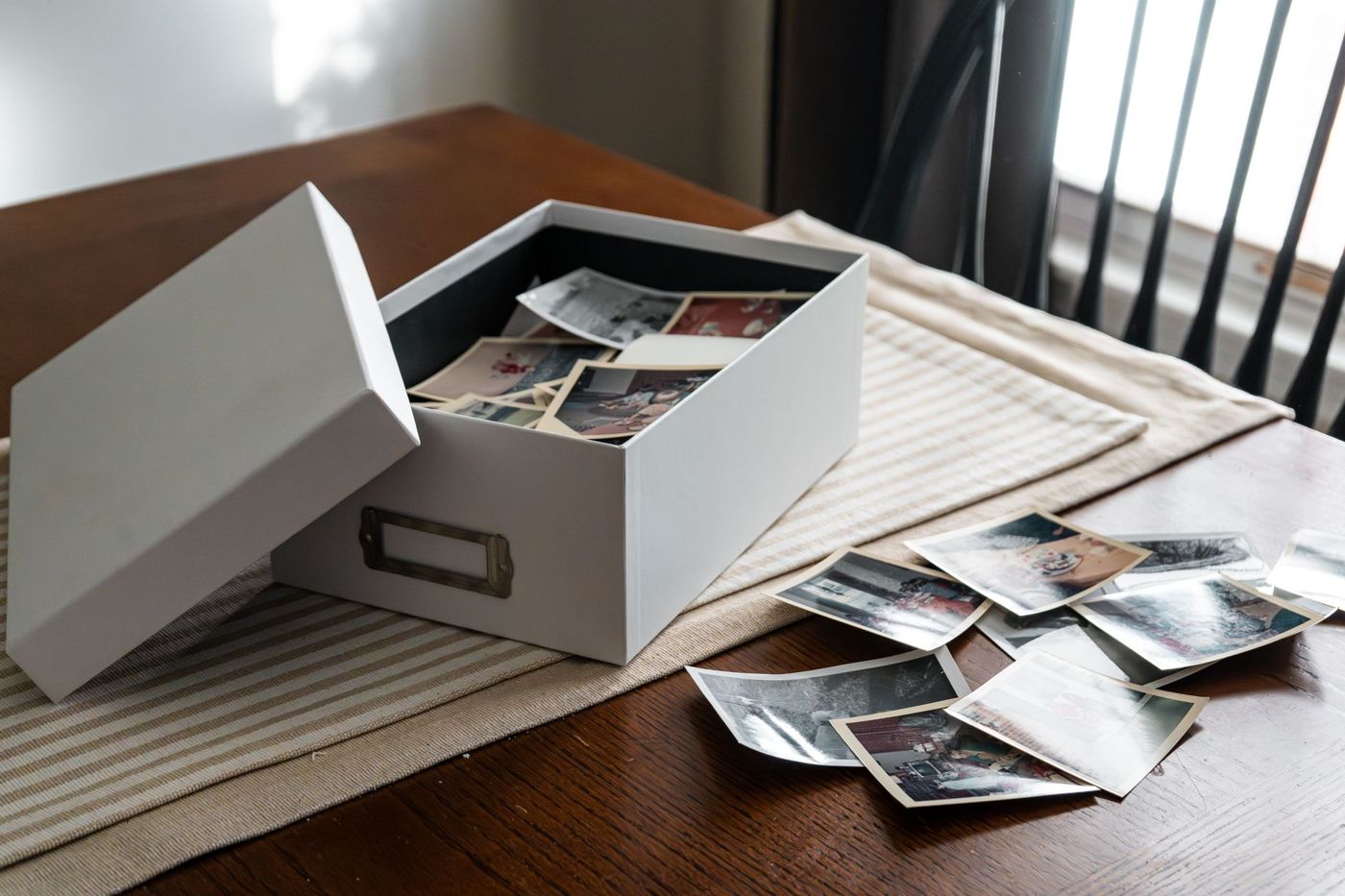 An open white photo box full of old colour prints on a wooden table, with loose photos beside it
