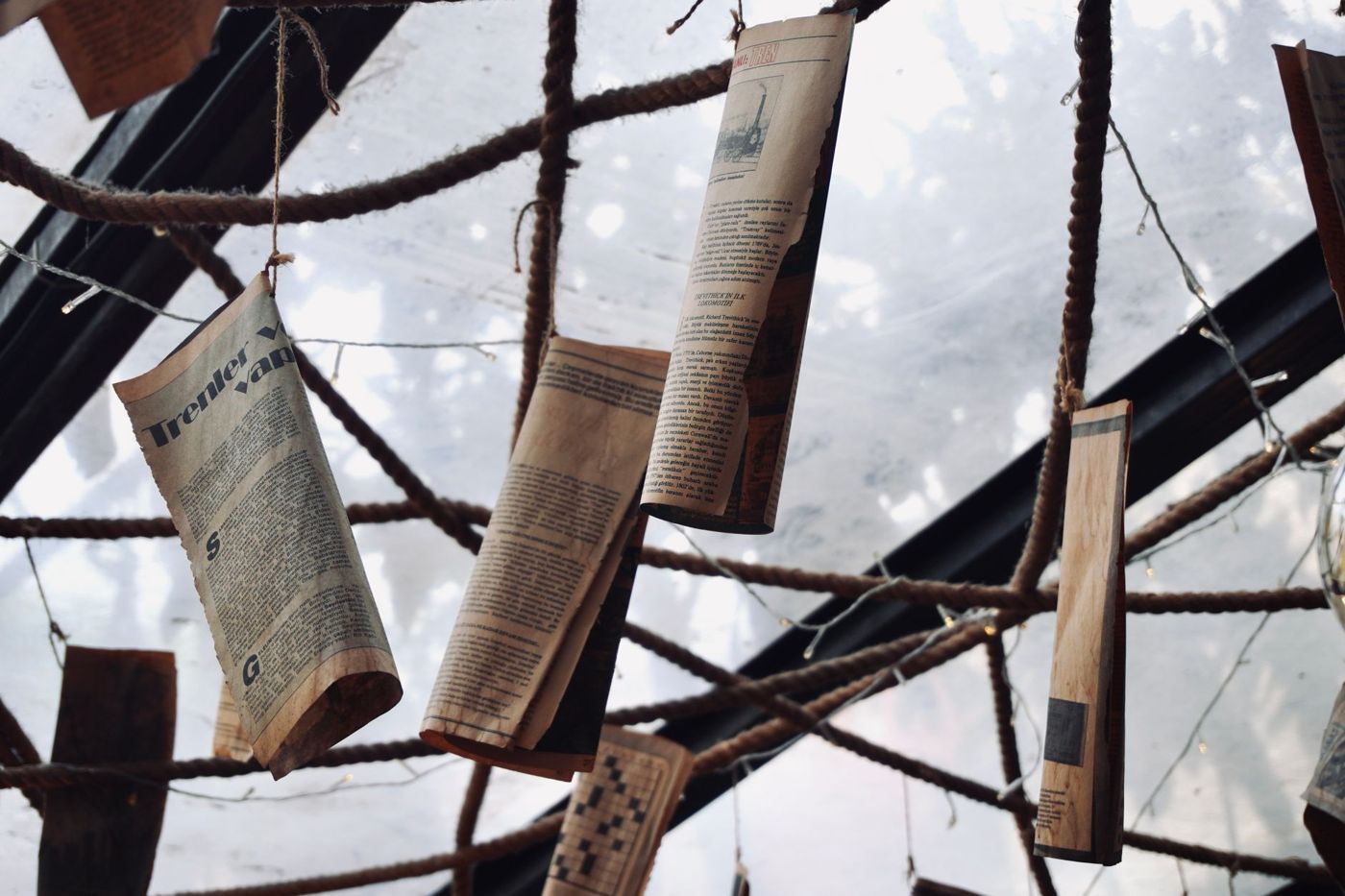 Newspaper cuttings with text hanging from ropes against a ceiling of rope and steel beams