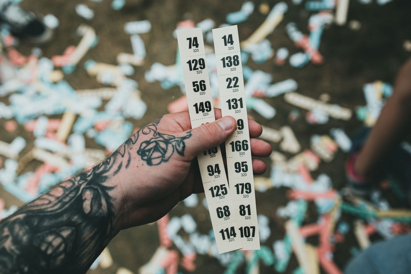 A tattooed hand holding two strips of numbered tickets above confetti on the ground