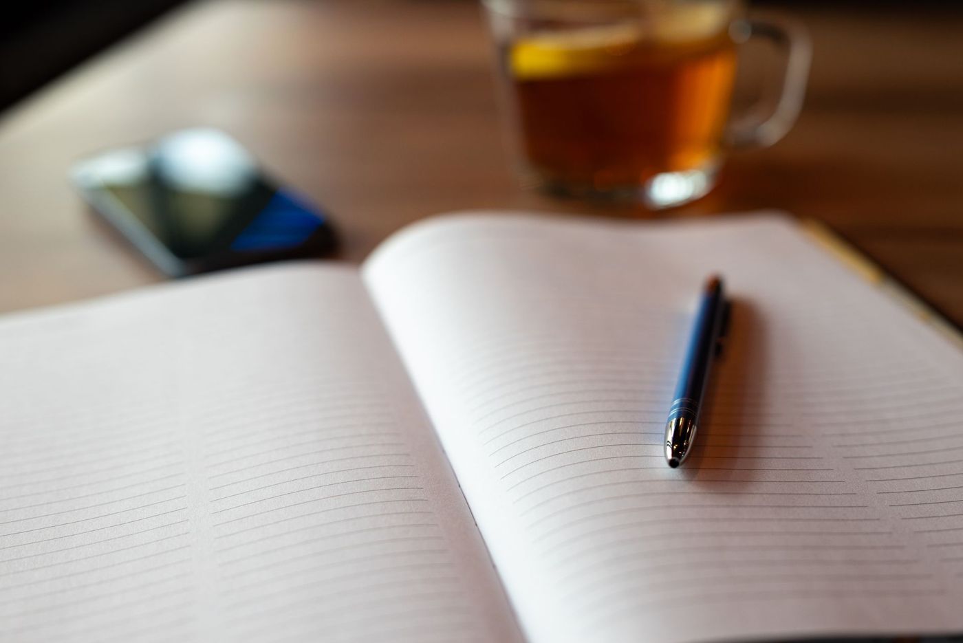 Quaderno a righe aperto con una penna blu, sullo sfondo sfocati un bicchiere di tè e uno smartphone su un tavolo di legno