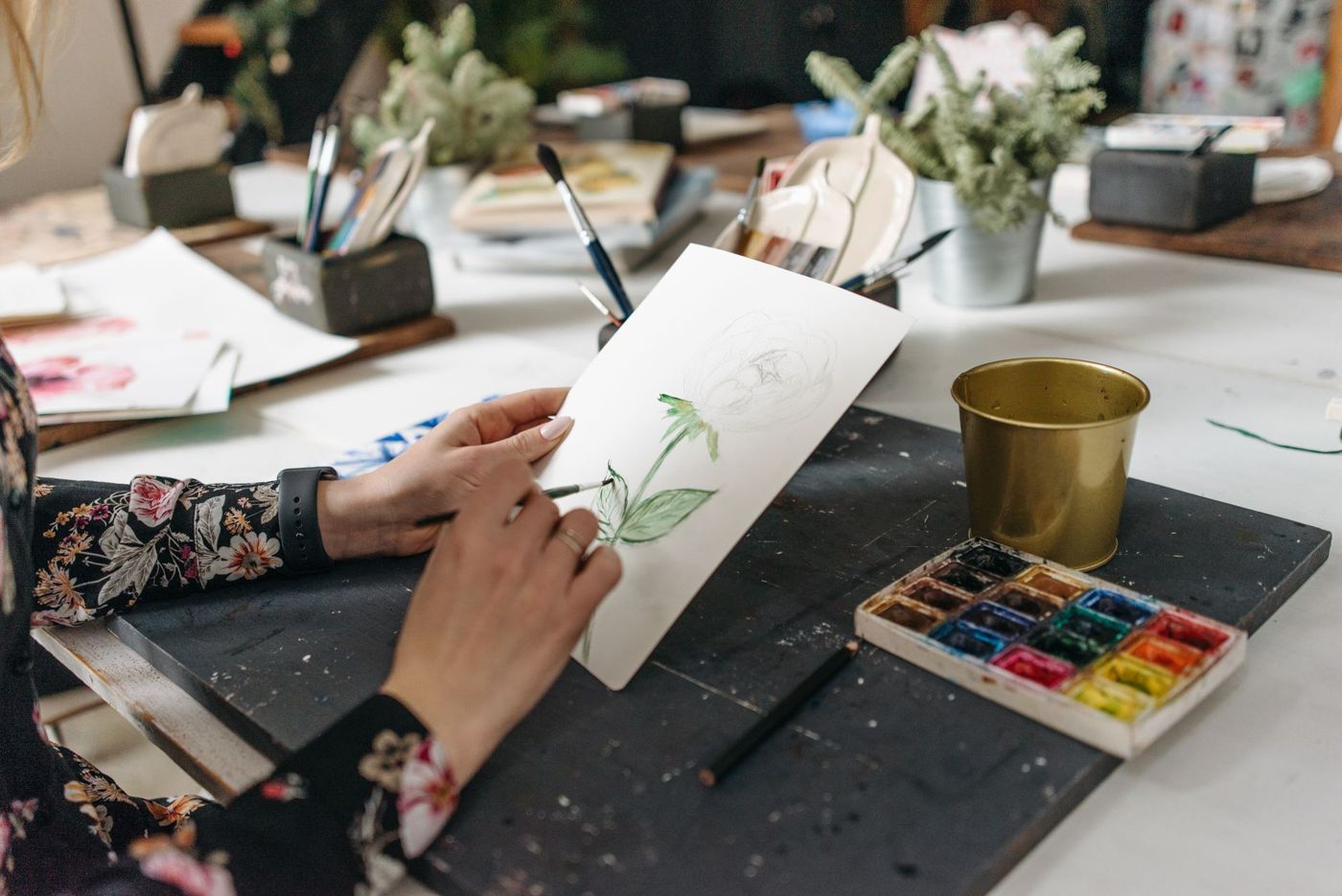 Two hands painting a green flower stem on a sheet of paper with a brush, next to a watercolour set and pots of brushes