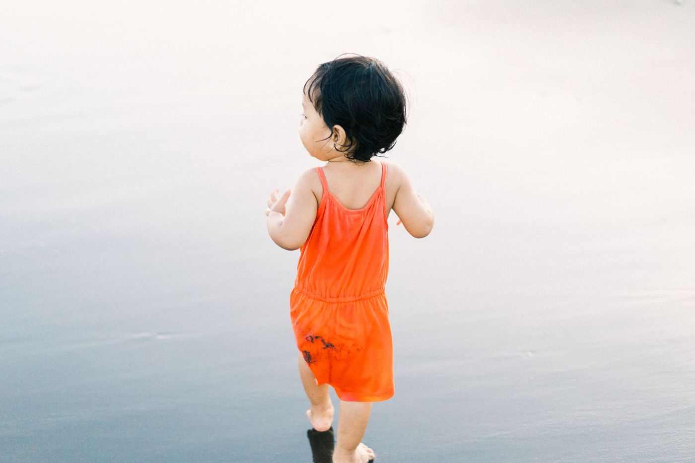 Toddler in an orange jumpsuit, seen from behind, walking across a calm, glistening beach