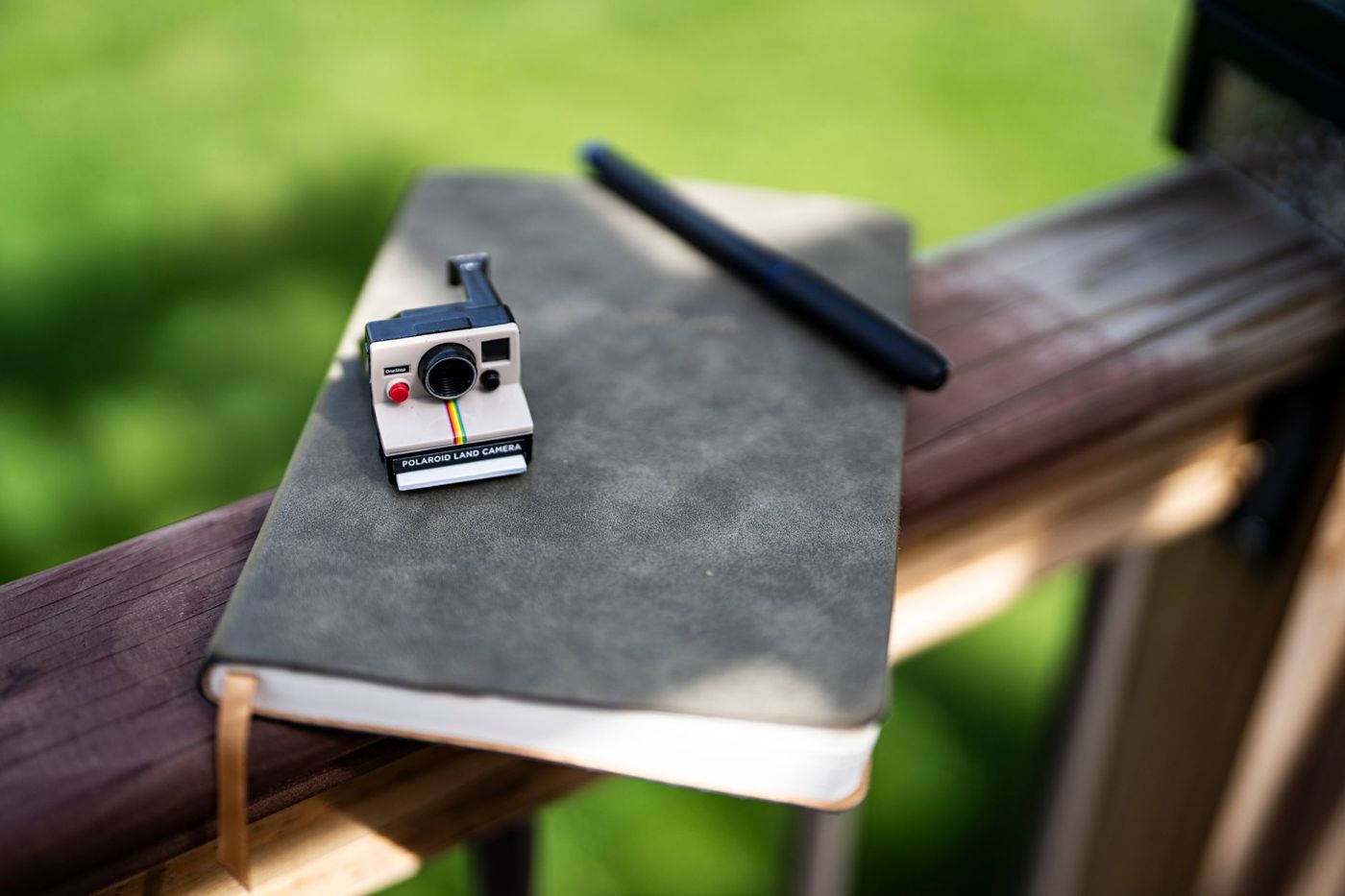 Malý model fotoaparátu Polaroid na šedém zápisníku s černou propiskou, na dřevěném zábradlí na slunci