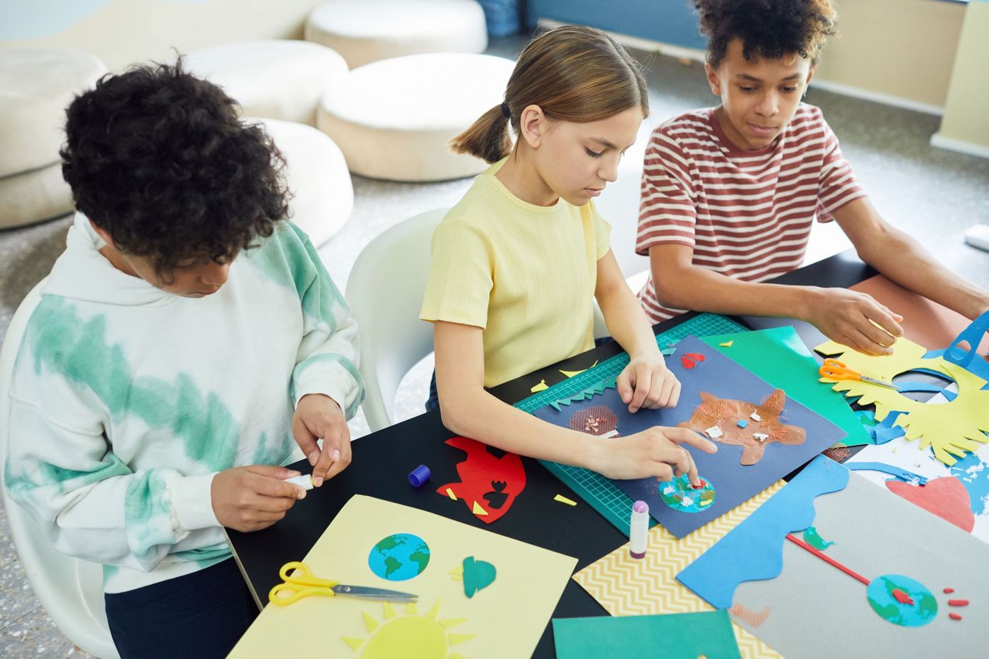 Tre bambini ritagliano e incollano a un tavolo con carta colorata, forbici e colla stick