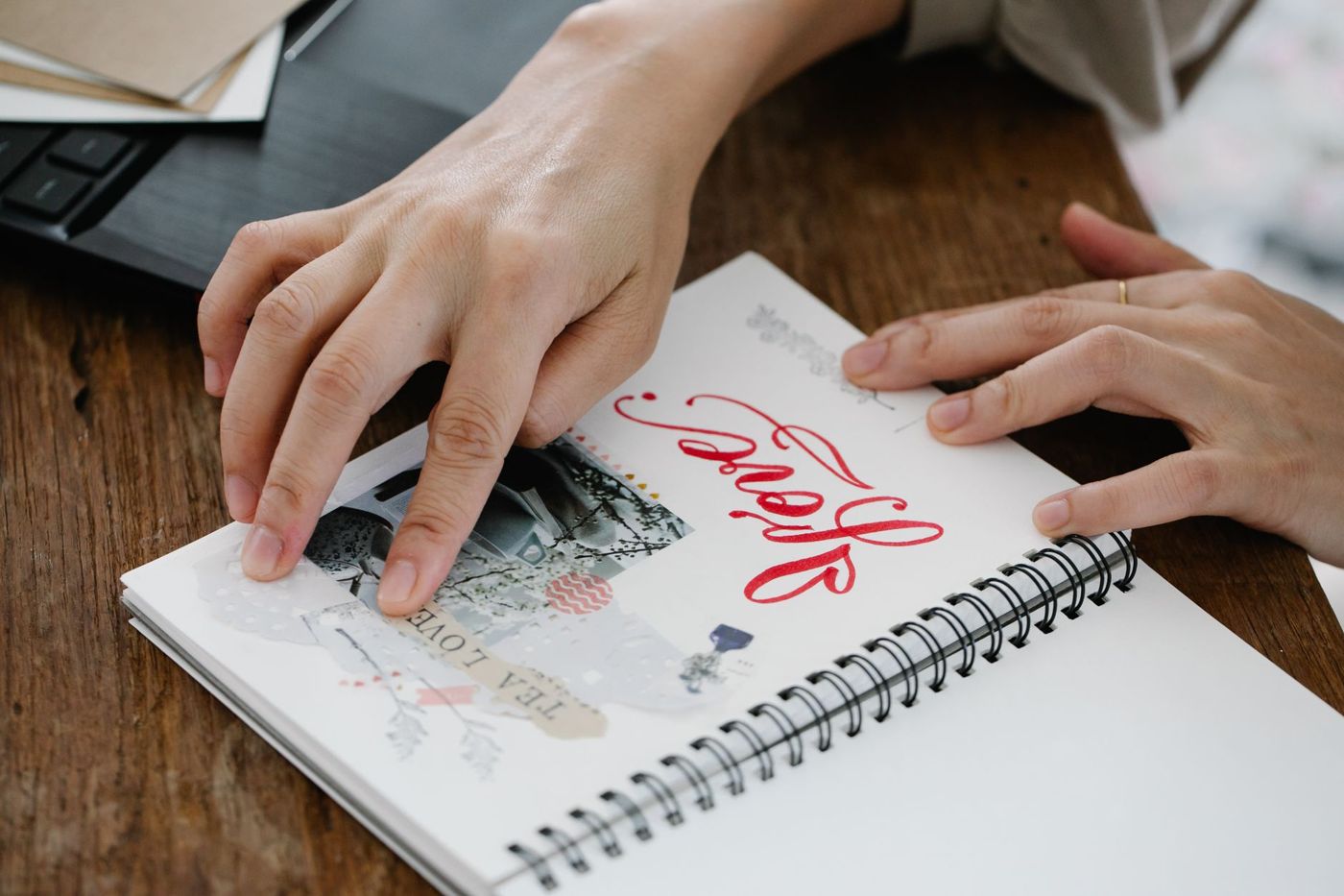 Two hands on an open spiral-bound notebook with a stuck-in photo, washi tape and red hand lettering