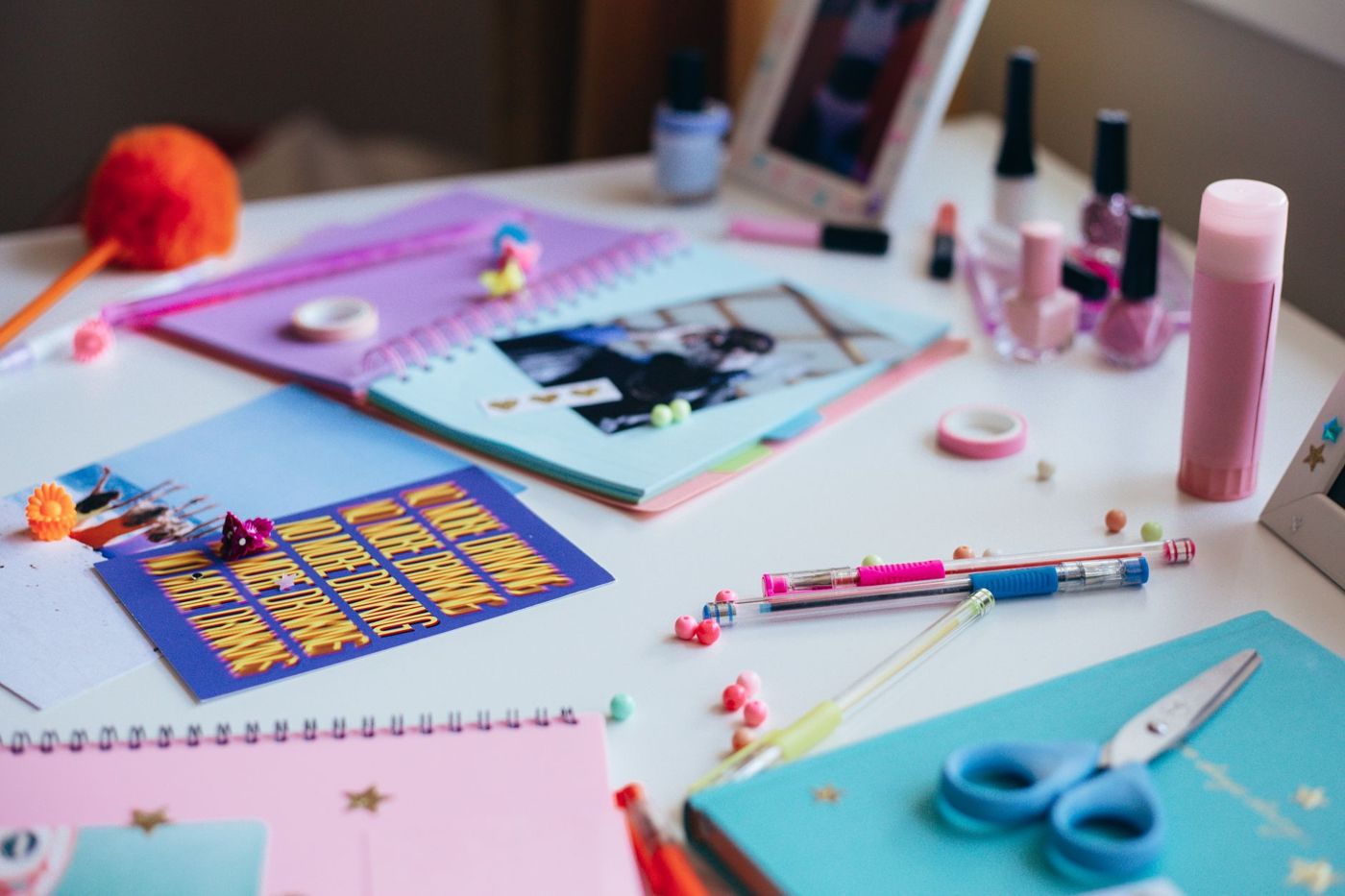 Craft table with colourful notebooks, a glue stick, gel pens, washi tape and scissors next to nail polish