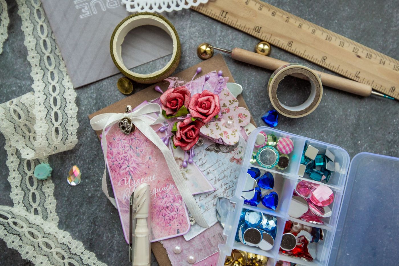 Scrapbooking supplies on a grey surface: lace, washi tape, rhinestones in a little box and a pink card with roses