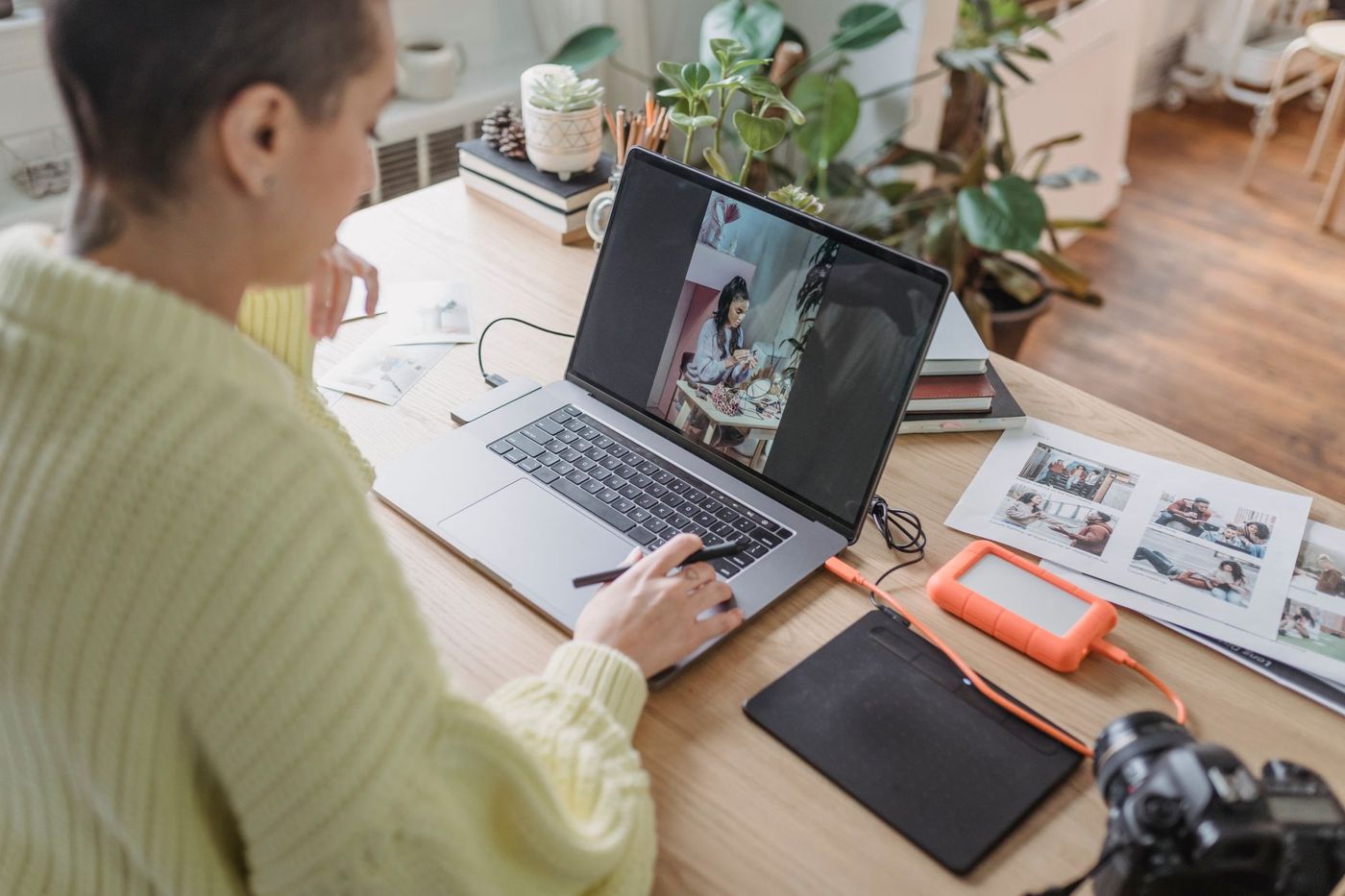Woman editing photos on a laptop with a drawing tablet, next to prints, an external drive and a camera