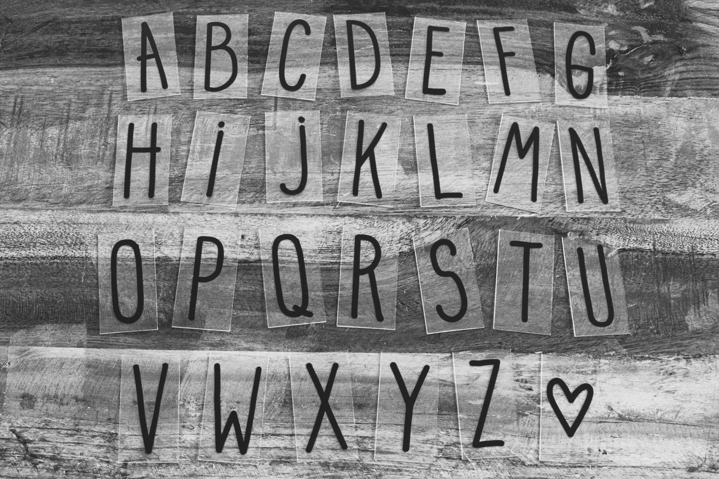 Black-and-white photo of the alphabet in black letters on clear strips on a wooden surface, with a little heart