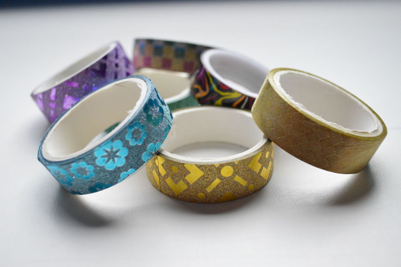 Six rolls of glittery and patterned washi tape in blue, gold and purple on a light background