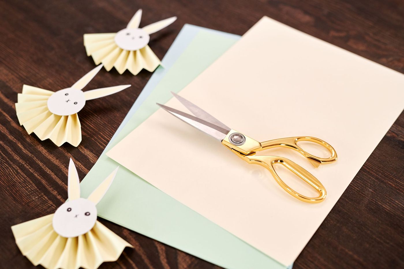 Gold scissors on sheets of cream, green and blue paper, next to three paper rosettes shaped like a little bunny