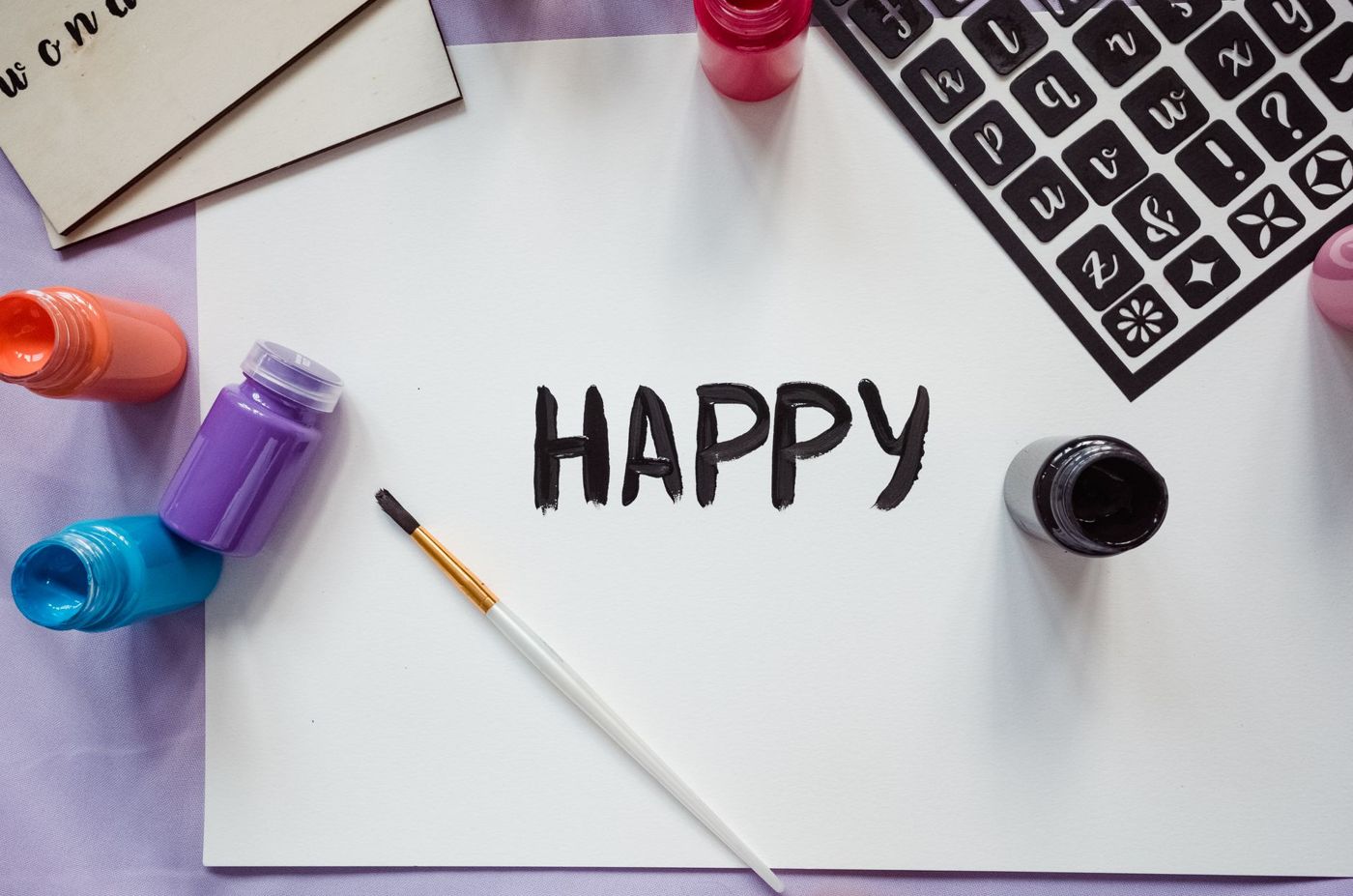 The word HAPPY in black paint on white paper, with a brush, pots of paint and a black lettering stencil beside it