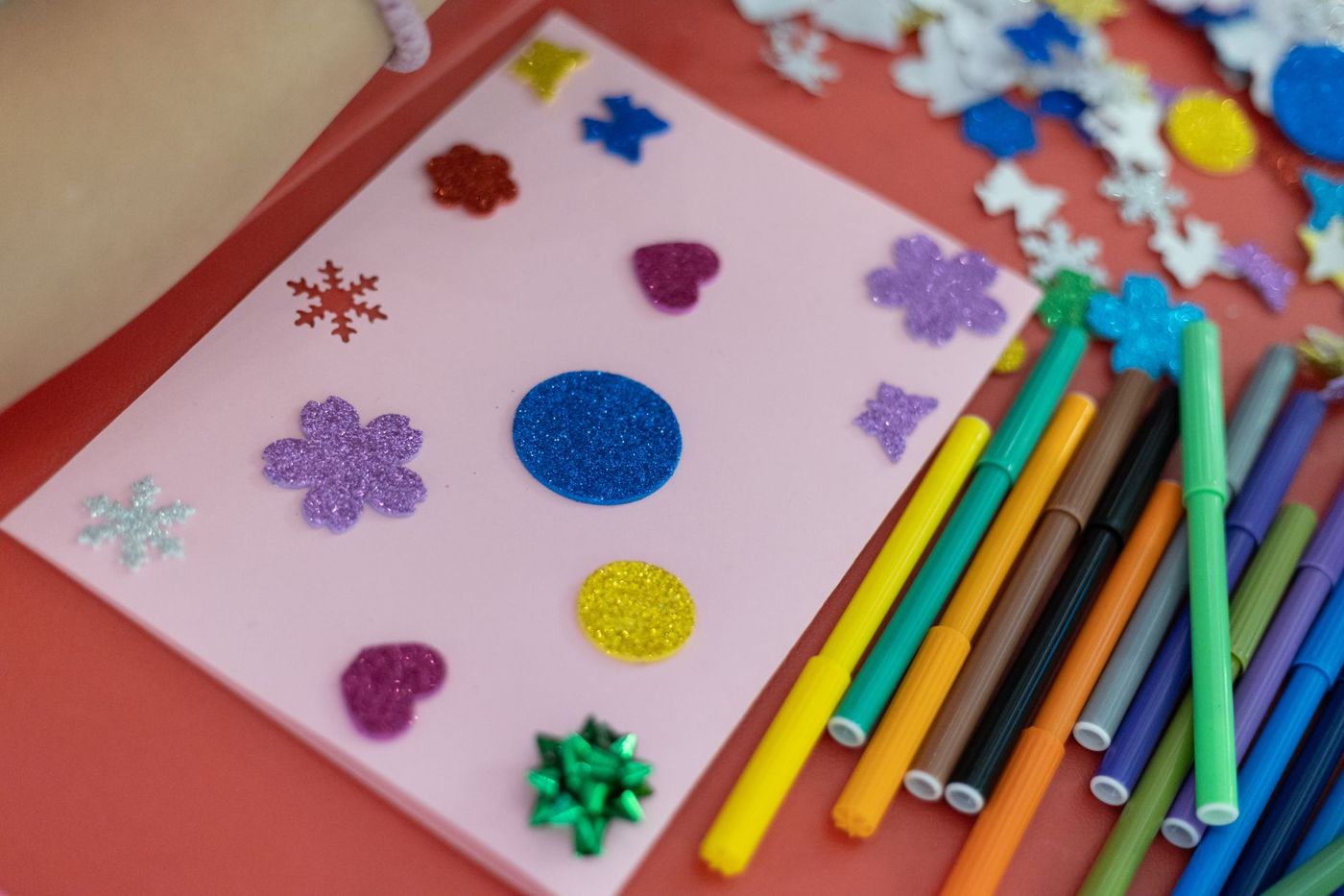 Pink card with glitter stickers shaped like hearts, flowers, snowflakes and circles, next to a fan of coloured markers