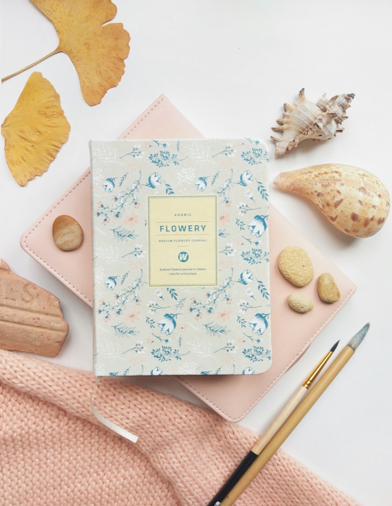 Notebook with a floral cover among seashells and a pen on a pink background